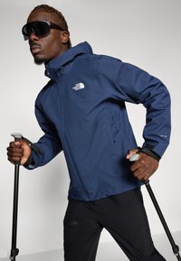 Marine vandtæt jakke med hætte, der har North Face-logo, lynlås-lommer og justerbare manchetter. Modellen holder på vandrestave.