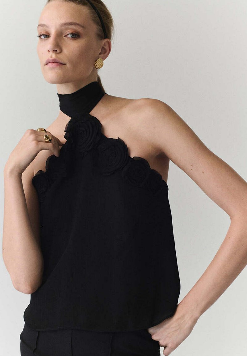 Femme portant un haut noir dos nu avec des détails de rose, des boucles d'oreilles dorées et des bagues, posant avec la main près du visage sur un fond uni.