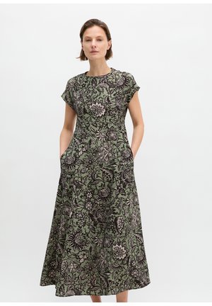 Kobieta w zielonej sukience midi z czarno-białym kwiatowym wzorem, krótkim rękawem i rękami w kieszeniach na jednolitym jasnym tle.