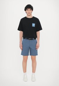 Tricou din bumbac negru cu detaliu albastru la buzunar, asortat cu pantaloni scurți albaștri și adidași albi. Tricoul are mâneci scurte și o croială lejeră.