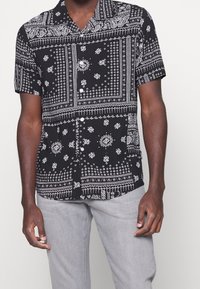 Chemise noire à manches courtes avec boutons, ornée de motifs complexes paisley et floraux blancs, dotée d'un col et d'une coupe décontractée. Associée à un pantalon gris clair.