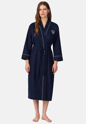 Vrouw draagt een marineblauwe knielange badjas met witte biezen, gestrikt in de taille, staand op blote voeten tegen een effen achtergrond.