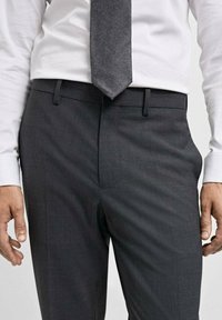 Sive patterned hlače glatke teksture, uparene s bijelom košuljom i tamnosivim kravatom, pokazuju moderne dizajnerske elemente i uredan kroj.