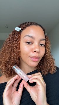 Heldere gloss lipproduct in een transparante tube met een zilveren dop. Model houdt de tube vast terwijl ze lichtbruin krullend haar en een natuurlijke huid toont.