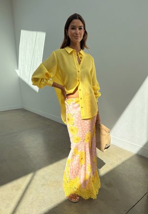 Γυναίκα που στέκεται στο φως του ήλιου, φορώντας ένα φαρδύ κίτρινο πουκάμισο και μια μακριά ροζ φούστα με κίτρινα λουλουδάτα μοτίβα, κρατώντας μια μπεζ τσάντα χειρός.