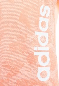 Sportovní top v broskvové barvě s texturovaným povrchem a výrazným bílým logem "adidas" na levé straně.