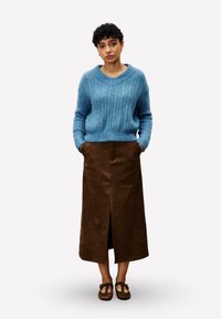 Femme aux cheveux courts et bouclés portant un pull en tricot bleu, une jupe midi marron avec une fente à l'avant et des chaussures T-strap marron, se tenant avec les mains dans les poches.