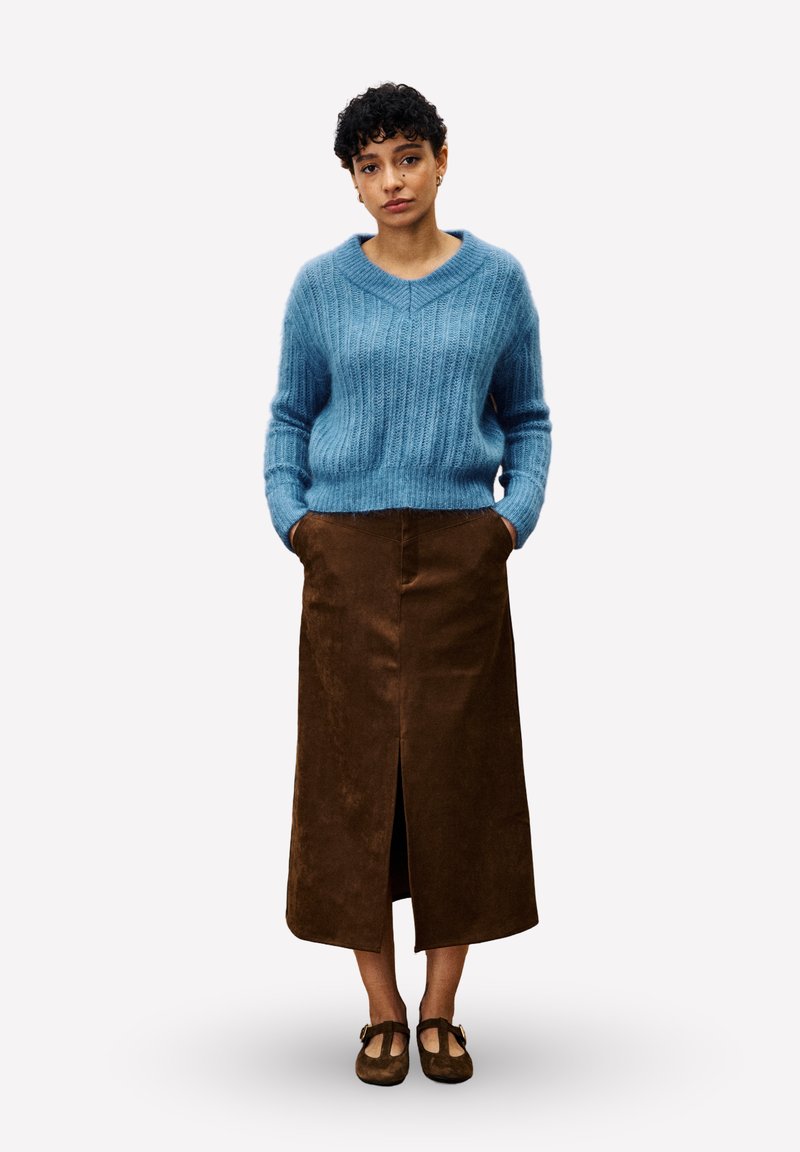 Femme aux cheveux courts et bouclés portant un pull en tricot bleu, une jupe midi marron avec une fente à l'avant et des chaussures T-strap marron, se tenant avec les mains dans les poches.