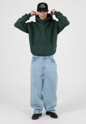 Sudadera verde con capucha y bolsillo frontal, combinada con vaqueros azul claro de gran tamaño. El modelo lleva zapatos negros y una gorra negra.