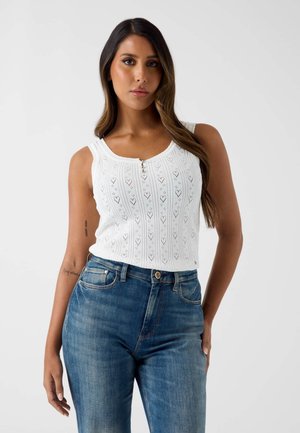 Frau mit langen Haaren, die ein ärmelloses weißes Strickoberteil mit Knöpfen und hoch taillierte blaue Jeans vor einem schlichten Hintergrund trägt.