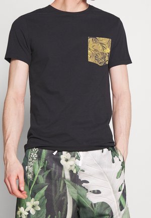 Homme portant un t-shirt noir à manches courtes avec une poche à imprimé tropical jaune et un short à motif floral vert, mains dans les poches.