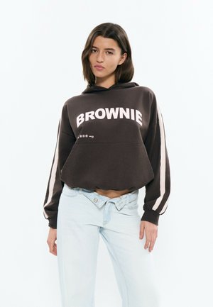 Joven con sudadera oscura con el texto "BROWNIE" y jeans azul claro con cintura asimétrica abotonada, de pie frente a un fondo blanco.