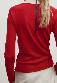 Haut rouge à manches longues avec un col rond et un détail de ruban noir à l'arrière. Texture lisse, coupe près du corps. Uni sans motifs.