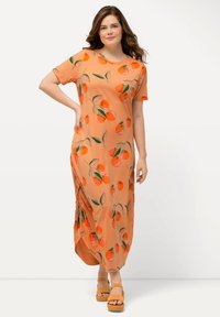 Orange maxi kjole med korte ærmer, prydet med et frugtmønster. Glat stof, sideslids for bevægelsesfrihed, bæres med tanfarvede platform sandaler.