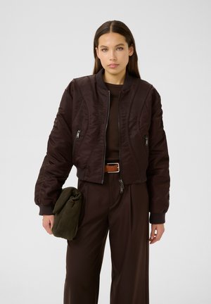 Brown bomber jacket with quilted texture, ribbed cuffs, and zippered pockets. Worn with brown trousers and an olive green bag.