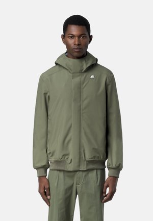 Uomo che indossa una giacca con cappuccio verde oliva e pantaloni coordinati, in piedi con le braccia rilassate contro uno sfondo chiaro e uniforme.