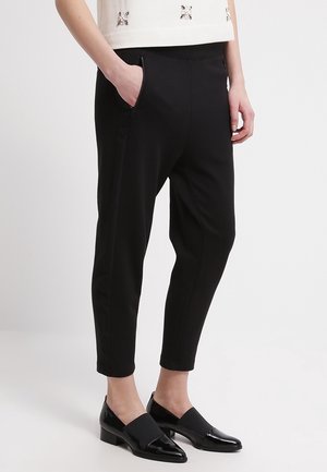 Person trägt schwarze, schmal zulaufende Hosen mit Seitentaschen und schwarze, lackierte Loafers mit niedrigem Absatz, steht vor einem schlichten weißen Hintergrund.