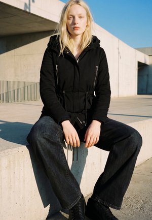 Młoda kobieta z długimi blond włosami, ubrana w czarną kurtkę i ciemne szerokie dżinsy, siedzi na betonowej krawędzi na świeżym powietrzu w świetle słonecznym.
