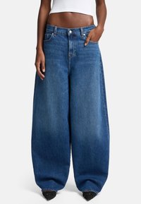 Jeans in denim blu oversized con un design a gamba larga, vita alta e cuciture visibili. Tessuto texturizzato con una finitura classica in lavaggio sbiadito.