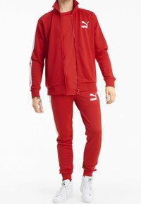 Tuta Puma rossa con giacca a zip e pantaloni coordinati. Presenta strisce bianche lungo le maniche e i pantaloni, logo sul petto e polsini a costine.