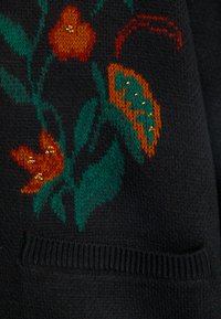 Juodas megztinis su gėlių raštu, kurį sudaro oranžinė ir žalia spalvos su nedideliais auksiniais akcentais. Įtraukta krašto rašto ir tekstūros detalė.