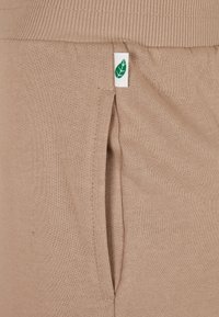 Bežinės medvilninės kelnės su elastingu liemeniu, šoninėmis kišenėmis ir mažyte žalia lapo logotipo etikete šone. Lygus pavircius.
