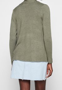 Oliivinvihreä neulepusero pitkillä hihoilla, ribbed-kauluksella ja sivurei'illä, käytetty vaaleansiniseksi farkkuhameen päällä, jossa on repaleinen helma.