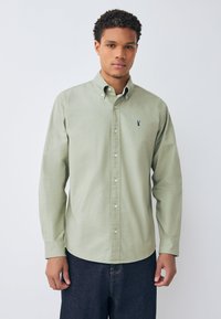 Camicia verde chiaro con bottoni e colletto, con un piccolo logo blu scuro sul petto e maniche lunghe. Indossata con jeans scuri.
