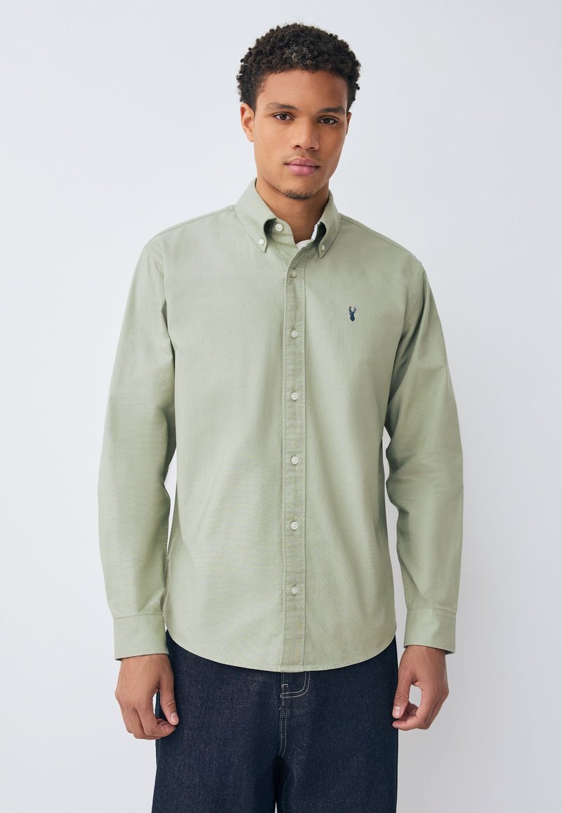 Camicia verde chiaro con bottoni e colletto, con un piccolo logo blu scuro sul petto e maniche lunghe. Indossata con jeans scuri.