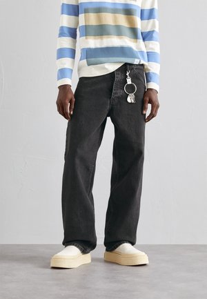 Person wearing black wide-leg jeans with a keychain and white slip-on shoes, paired with a blue, white, and beige striped long-sleeve top.