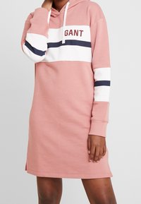 Robe sweat à capuche rose avec des rayures blanches et bleu marine, arborant un logo sur la poitrine et des poignets côtelés. Des détails avec fentes latérales sont visibles.