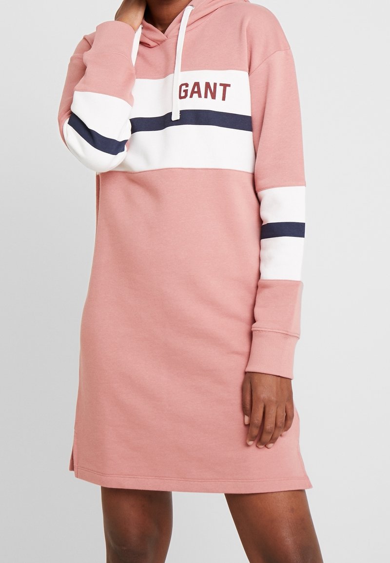 Robe sweat à capuche rose avec des rayures blanches et bleu marine, arborant un logo sur la poitrine et des poignets côtelés. Des détails avec fentes latérales sont visibles.