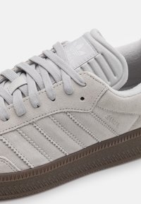 Szürke velúr sneaker, textúrált felülettel, három csíkos díszítéssel, kerek fűzőkkel és bordázott sarkal. Logó a nyelvén és az oldalán.