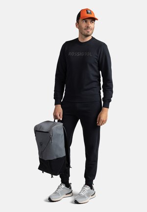 Grau und schwarzer Rucksack mit Reißverschluss, schwarzes Sweatshirt mit Logo, schwarze Jogginghose, graue Sneakers und eine orange-schwarze Mütze.