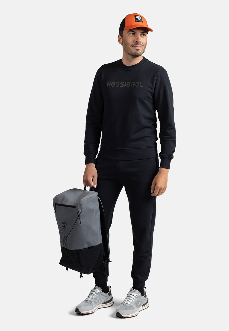 Zaino grigio e nero con zip, felpa nera con logo, pantaloni felpati neri, sneakers grigie e berretto arancione e nero.