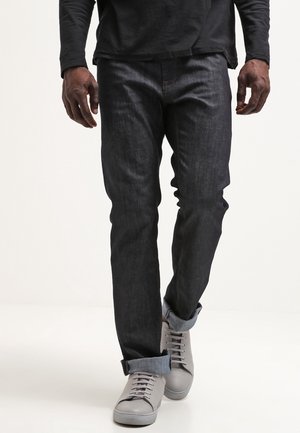 Mann trägt dunkle hochgekrempelte Jeans, graue Turnschuhe und ein schwarzes Langarmshirt und geht vor einem schlichten weißen Hintergrund.