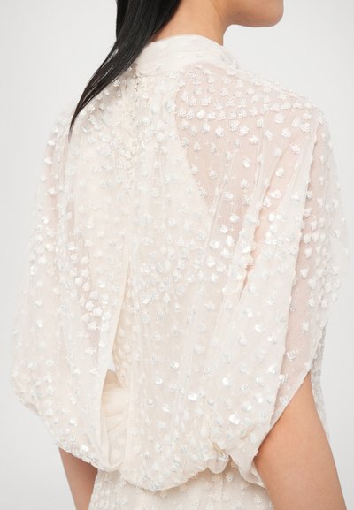 Priehľadná krémová tkanina so striebornými flitrami vytvárajúcimi bodkovaný vzor; voľne padajúce rukávy a vysoký golier vzadu.