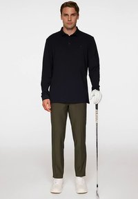 Schwarzes Langarm-Poloshirt, olivgrüne Hose, weiße Turnschuhe und ein Golfschläger. Das Polo hat einen Kragen und ein dezentes Branding auf der Brust.