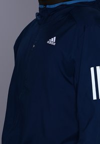 Chaqueta cortavientos azul marino con cierre frontal, capucha y logo de Adidas en blanco en el pecho. Cuenta con tres rayas blancas en el brazo izquierdo.