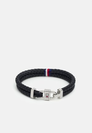Pulsera trenzada de cuero negro con cierre plateado que presenta un acento y detalle de logo en rojo, blanco y azul.