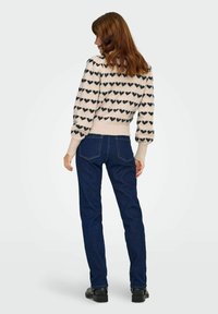 Jerséi de punto en color crema con patrones de corazones negros, puños y bajo acanalados, combinado con jeans azul oscuro de pierna recta, calzado con zapatos negros.