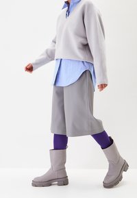 Suéter gris de manga larga con cuello en v sobre una camisa de rayas azules, combinado con pantalones cortos grises, leggings morados y botines grises con suelas gruesas.