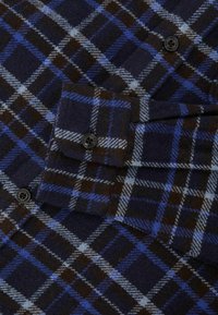 Camicia di flanella con motivo a quadri blu navy e marrone con accenti azzurri. Bottoni neri sulle tasche e sui polsini. Texture morbida.