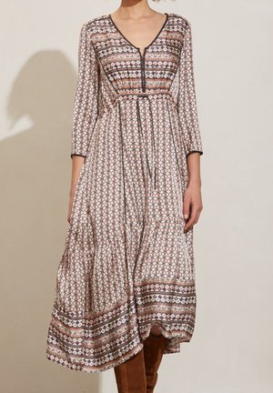 Femme portant une longue robe bohème à motifs avec des manches trois-quarts et des bottes marron montant jusqu'aux genoux sur un fond uni.