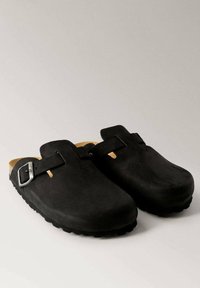 Black suede clogs with an open back and a single adjustable buckle. Textured rubber sole for grip. Inner lining in a tan color.