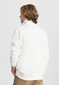 Felpa bianca in fleece con colletto alto e polsini a coste, dal taglio morbido e dalla texture soffice. Dettagli di cucitura visibili alle cuciture.