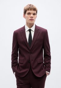 Μπορντό κοστούμι με εφαρμοστή γραμμή, που διαθέτει μονόκλειστο σχέδιο, δύο μπροστινές τσέπες, μαύρα κουμπιά και λευκό πουκάμισο με μαύρη γραβάτα.