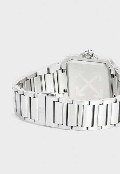 Montre bracelet en métal argenté avec un fond de boîtier carré et un motif de flèches gravé au dos et sur la boucle, vue sous un angle arrière.
