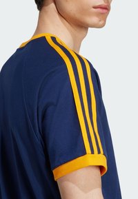 Navy blue t-shirt with yellow striped accents on sleeves and collar. Made from cotton, featuring a round neckline and soft texture.