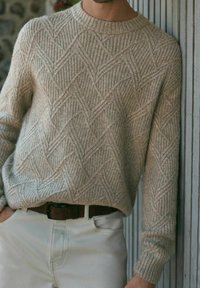 Sweter dzianinowy w jasnym beżu, z teksturowanym wzorem w romby. Długie rękawy, okrągły dekolt i luźny krój. Połączony z białymi spodniami.